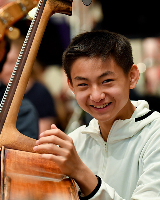 An Asian male RCMJD student, holding his cello, smiling at the camera, sitting 